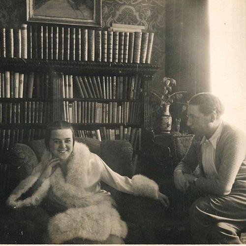 A black and white photograph of Kitty sitting in a fur trimmed coat, with Alfred sitting to the right looking at her.