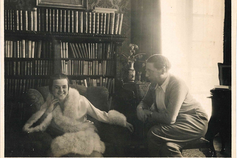A black and white photograph of Kitty sitting in a fur trimmed coat, with Alfred sitting to the right looking at her.