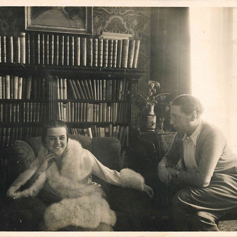 A black and white photograph of Kitty sitting in a fur trimmed coat, with Alfred sitting to the right looking at her.