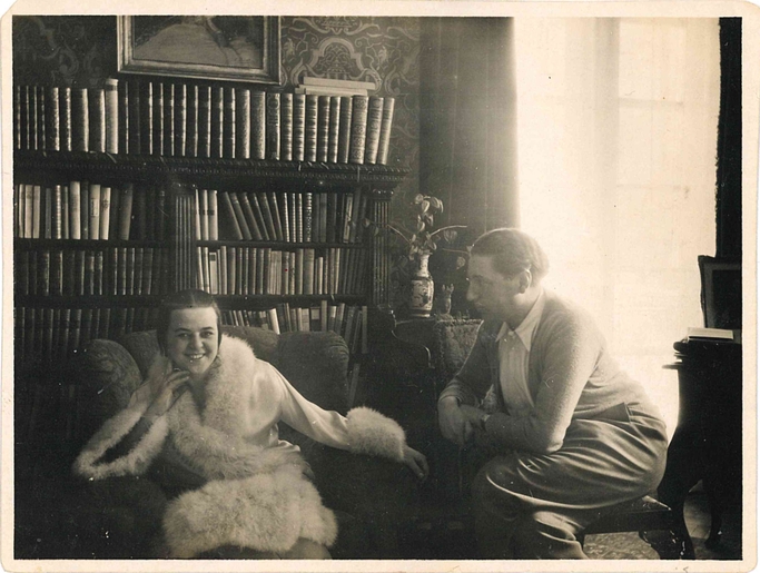 A black and white photograph of Kitty sitting in a fur trimmed coat, with Alfred sitting to the right looking at her.