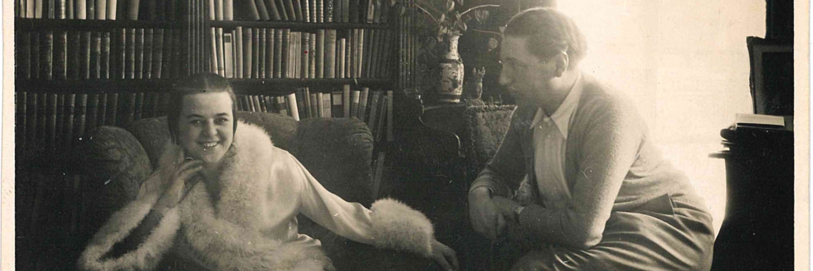 A black and white photograph of Kitty sitting in a fur trimmed coat, with Alfred sitting to the right looking at her.